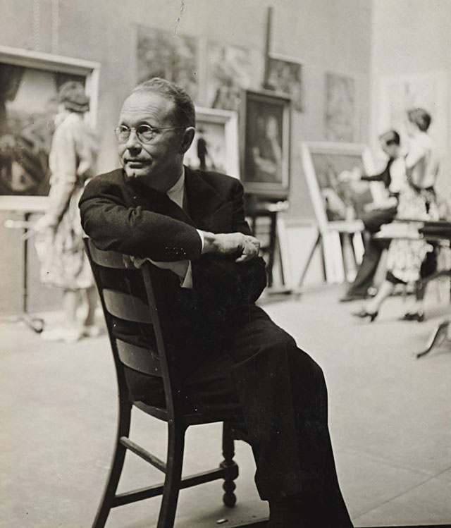 Artist Grant Wood seated in a gallery looking away from the camera with three people viewing pieces of art on display in the background.