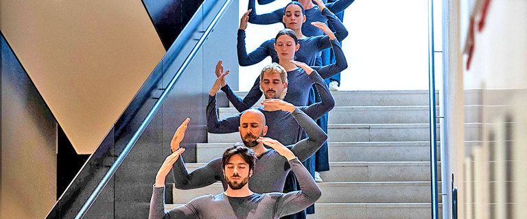 6 people wearing grey hold their arms over their heads while standing on stairs