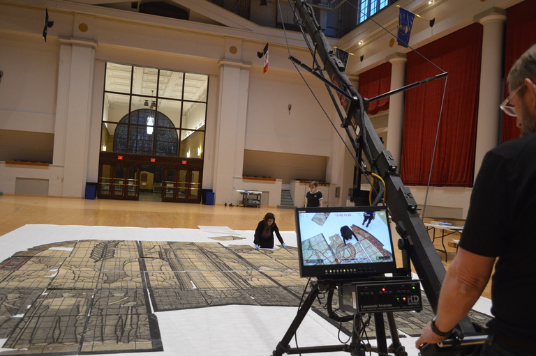 woman kneels with a pieces of a drawing of a stained glass window while a man operates of boom camera