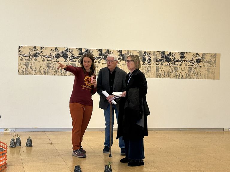 a woman points while two other people look on in front of a long print on a white wall