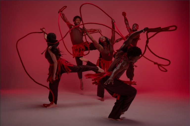 5 people in brown clothes whip red ropes in front of a pale red backdrop