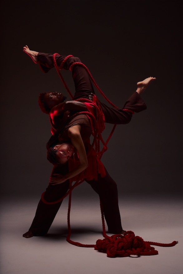 A black male performer lifts a black female performer upside down, twisted in red rope against a dark gray background 