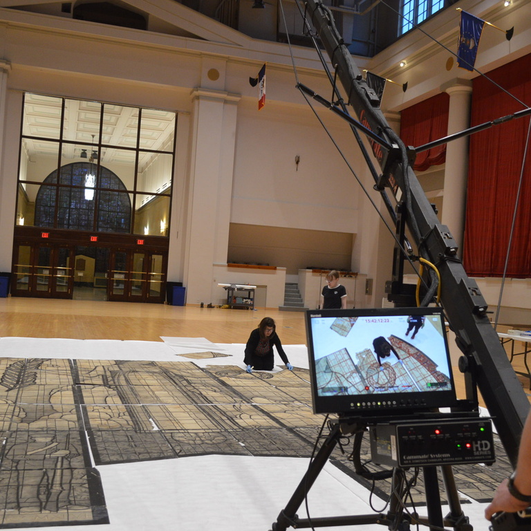 woman kneels with a pieces of a drawing of a stained glass window while a man operates of boom camera